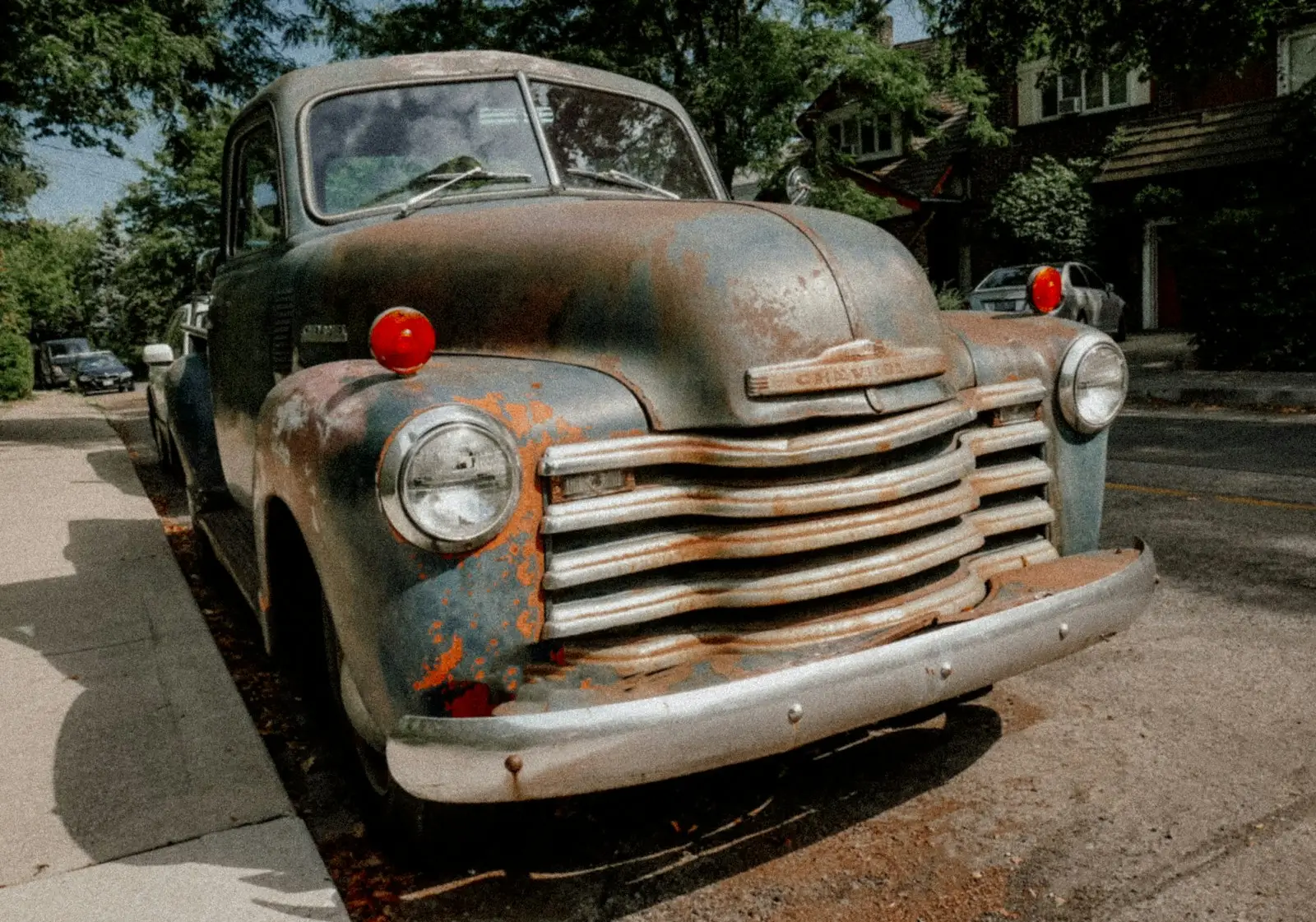 A rusted old Chevy pickup parked at a curb on a tree-lined street, in black and white